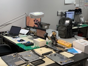 A workbench is cluttered with technology repair tools and disassembled electronic devices. Multiple laptops are open, and an overhead lamp provides focused lighting on the workspace. A backpack sits at the back, amidst a disarray of tools such as screwdrivers, a utility knife, and an organized box of small components. Various pieces of electronic hardware and circuit boards are spread out on the table.