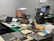 A workbench is cluttered with technology repair tools and disassembled electronic devices. Multiple laptops are open, and an overhead lamp provides focused lighting on the workspace. A backpack sits at the back, amidst a disarray of tools such as screwdrivers, a utility knife, and an organized box of small components. Various pieces of electronic hardware and circuit boards are spread out on the table.