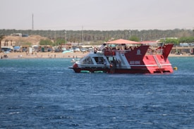 A red and white boat named 'Jules Verne' is sailing on a body of water. The boat is relatively large and populated with people on the upper deck, enjoying the sunny day. In the background, a sandy beach is visible, populated with several parked vehicles, small beach huts, and beachgoers. The shoreline is lined with palm trees and the overall setting appears to be a coastal area on a bright, clear day.