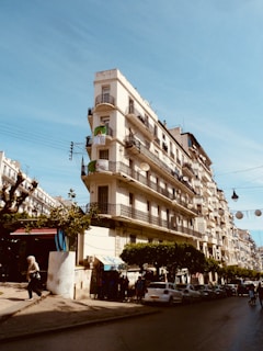 A multi-story building with a classic architectural style, featuring balconies and iron railings, is located on a street corner lined with trees. People are walking on the sidewalk, and cars are parked along the street. The Algerian flag is visible on one of the balconies against a clear blue sky.