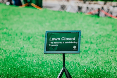 A green lawn with a sign that reads 'Lawn Closed. The new sod is establishing its roots.' The sign is positioned in the forefront on a metal stand, surrounded by vibrant green grass.