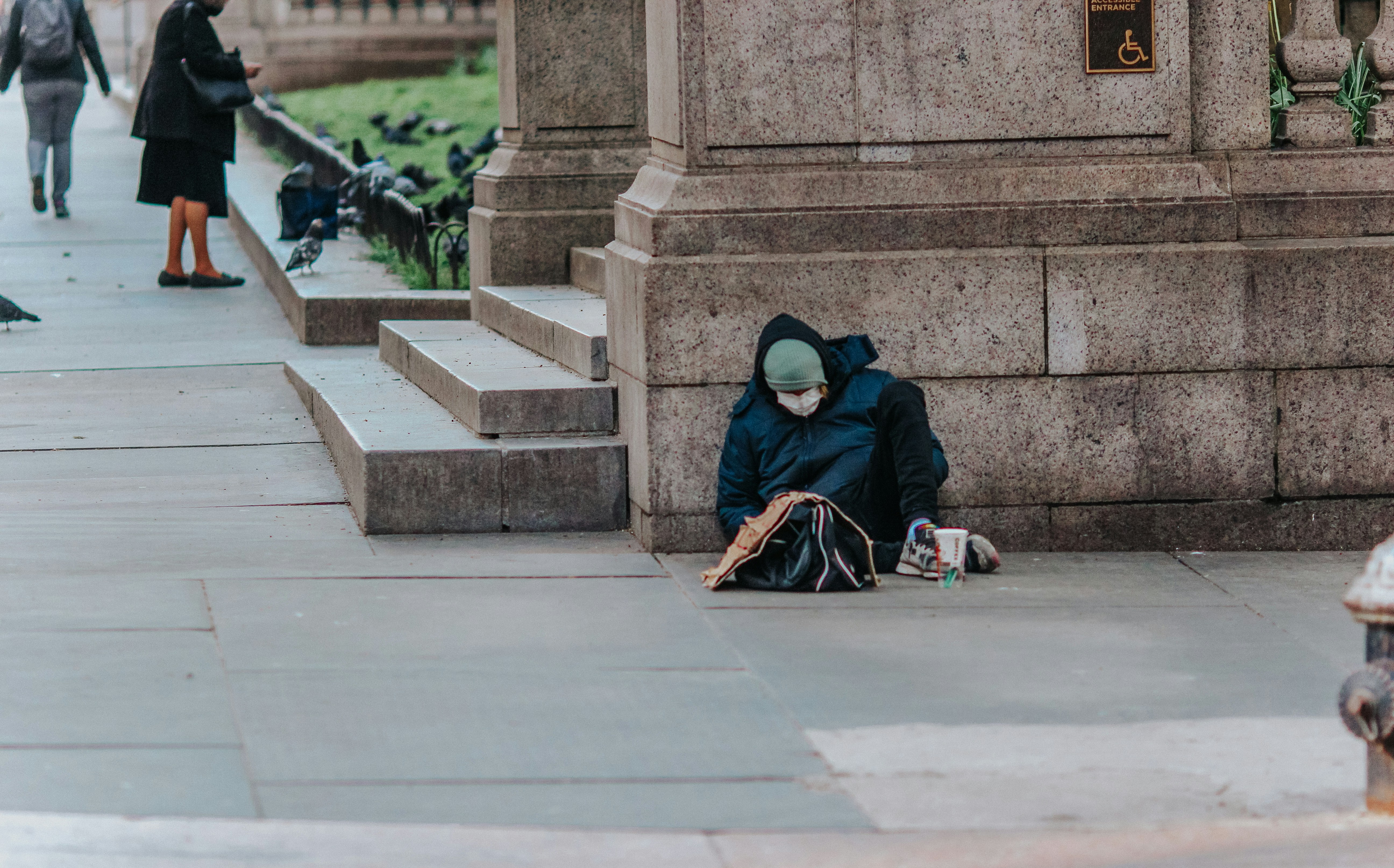 Homeless man sitting against concrete wall wearing a jacket, beanie and mask. In front of him are a bag and a cup.