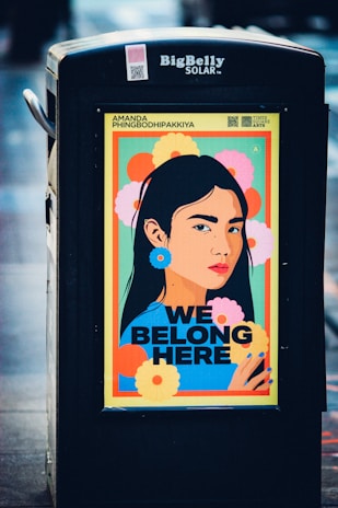 A brightly colored poster displayed on a public garbage bin features an illustrated portrait of a woman with long black hair. The background is filled with large, vibrant flowers in shades of yellow, orange, pink, and green. The text 'WE BELONG HERE' is prominently displayed in bold black letters below the portrait. The top of the poster includes the name 'Amanda Phingbodhipakkiya' and a QR code. The garbage bin has the label 'BigBelly SOLAR' at the top.