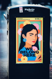 A brightly colored poster displayed on a public garbage bin features an illustrated portrait of a woman with long black hair. The background is filled with large, vibrant flowers in shades of yellow, orange, pink, and green. The text 'WE BELONG HERE' is prominently displayed in bold black letters below the portrait. The top of the poster includes the name 'Amanda Phingbodhipakkiya' and a QR code. The garbage bin has the label 'BigBelly SOLAR' at the top.