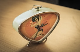 A vintage triangular alarm clock with a metallic frame and a retro-style illustration on the clock face, depicting a pin-up girl. The numbers and hands of the clock are visible against an orange background. It rests on a wooden surface.