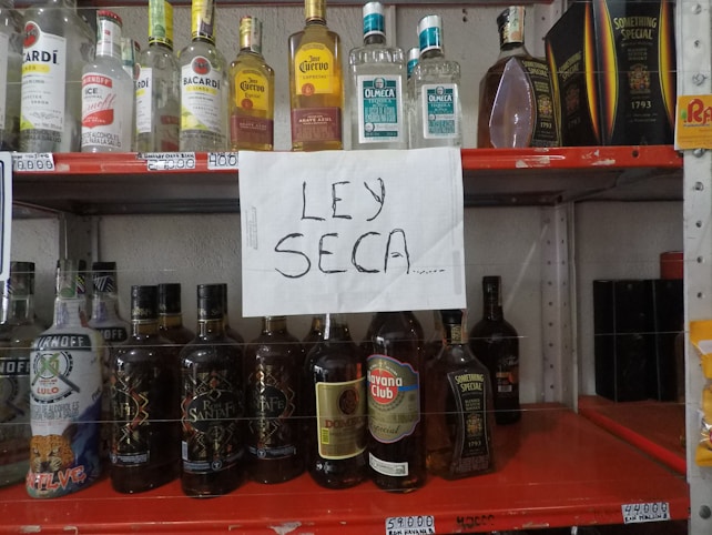 A shelf with various brands of alcoholic beverages, including bottles of vodka, rum, tequila, and whiskey. In the center, a handwritten sign reads 'LEY SECA' indicating a dry law or prohibition.