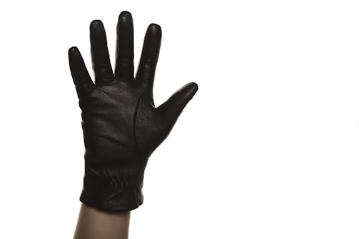 A black leather glove on a hand raised against a plain white background. The glove appears sleek and well-fitted, with its fingers spread open, emphasizing the texture of the leather.