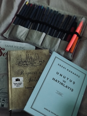 A collection of books and writing tools on a surface, including two visible book covers with text in a non-English script, and a set of pens neatly arranged in a fabric holder. The surrounding fabric has a soft, muted color.