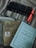 A collection of books and writing tools on a surface, including two visible book covers with text in a non-English script, and a set of pens neatly arranged in a fabric holder. The surrounding fabric has a soft, muted color.