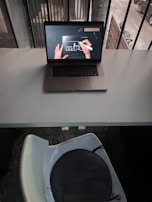A laptop is placed on a white desk, displaying a digital drawing or design on its screen with a person's hands using a stylus. The scene is indoors next to a window with vertical bars and there is a view of a street outside. Below the desk, there is a white chair with a dark circular object, possibly a bag, on it.