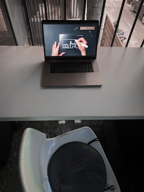 A laptop is placed on a white desk, displaying a digital drawing or design on its screen with a person's hands using a stylus. The scene is indoors next to a window with vertical bars and there is a view of a street outside. Below the desk, there is a white chair with a dark circular object, possibly a bag, on it.