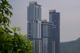 A modern high-rise building with a sleek glass facade, featuring multiple levels and large windows. It stands prominently against a hazy sky, with leafy green branches partially framing the foreground. The background includes distant hills, adding to the serene urban landscape.