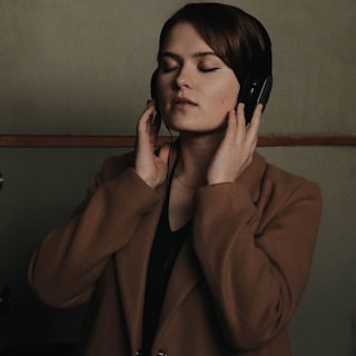 A person with closed eyes is enjoying music through headphones. They are wearing a brown coat and have a calm expression. The background is muted and plain, focusing attention on the subject.