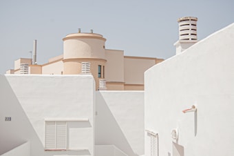 A minimalist architectural scene featuring a sequence of white and cream-colored buildings. The structures display clean lines and geometric shapes, including cylindrical vents or chimneys. The clear blue sky provides a serene backdrop to the simple, uncluttered urban setting.