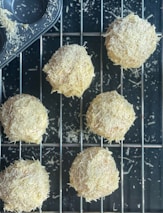 Freshly baked round pastries covered in shredded cheese rest on a cooling rack. A muffin tray with cheese remnants can be seen in the corner, suggesting the baking process. The scene depicts a rustic, homemade culinary creation.