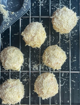 Freshly baked round pastries covered in shredded cheese rest on a cooling rack. A muffin tray with cheese remnants can be seen in the corner, suggesting the baking process. The scene depicts a rustic, homemade culinary creation.
