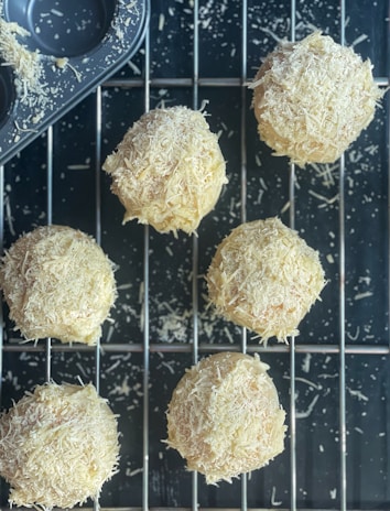 Freshly baked round pastries covered in shredded cheese rest on a cooling rack. A muffin tray with cheese remnants can be seen in the corner, suggesting the baking process. The scene depicts a rustic, homemade culinary creation.