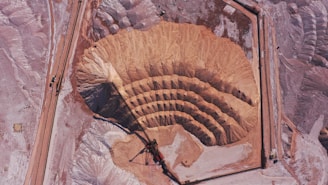 An aerial view of a large open-pit mine featuring concentric terraces carved into the earth, surrounded by barren, dusty terrain. There is heavy machinery at the bottom of the pit indicating active mining operations. The landscape is expansive and marked by shades of brown and pink.