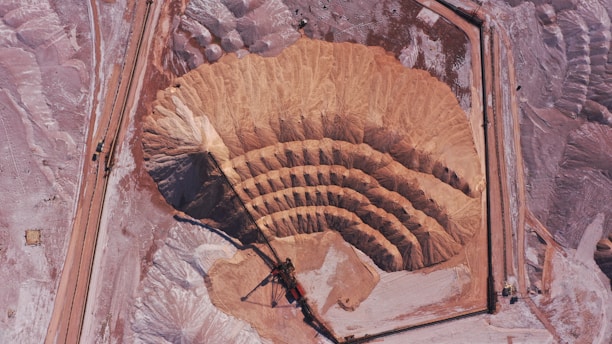 An aerial view of a large open-pit mine featuring concentric terraces carved into the earth, surrounded by barren, dusty terrain. There is heavy machinery at the bottom of the pit indicating active mining operations. The landscape is expansive and marked by shades of brown and pink.
