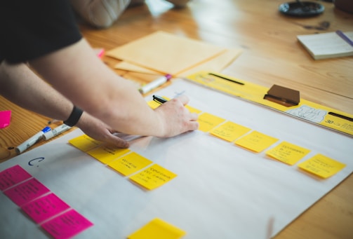 A person is organizing colorful sticky notes on a large sheet of paper placed on a wooden table. Several sticky notes are yellow, while a few are pink, with writings visible on them. There are markers and other stationery items around the paper.