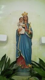 A statue of a woman wearing a blue robe and a golden crown, holding a child. The child is dressed in a white garment and holds a small globe. Both figures have serene expressions. The backdrop is a pale yellow wall, and there are green leaves at the base of the statue.
