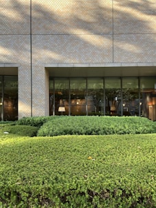 A building exterior with a facade made of light-colored bricks. Large, floor-to-ceiling glass windows line the wall, revealing an interior space that appears to be well-lit and furnished with elements like a lamp and framed art. In front of the building is a well-maintained garden with neatly trimmed bushes and greenery. Shadows from nearby trees are cast on the facade.