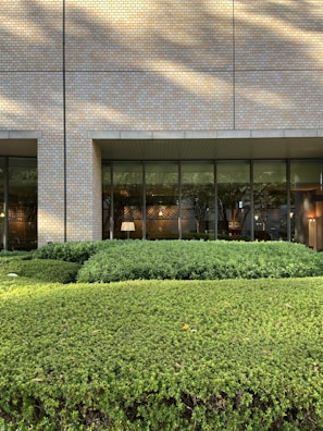 A building exterior with a facade made of light-colored bricks. Large, floor-to-ceiling glass windows line the wall, revealing an interior space that appears to be well-lit and furnished with elements like a lamp and framed art. In front of the building is a well-maintained garden with neatly trimmed bushes and greenery. Shadows from nearby trees are cast on the facade.