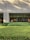 A building exterior with a facade made of light-colored bricks. Large, floor-to-ceiling glass windows line the wall, revealing an interior space that appears to be well-lit and furnished with elements like a lamp and framed art. In front of the building is a well-maintained garden with neatly trimmed bushes and greenery. Shadows from nearby trees are cast on the facade.