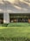 A building exterior with a facade made of light-colored bricks. Large, floor-to-ceiling glass windows line the wall, revealing an interior space that appears to be well-lit and furnished with elements like a lamp and framed art. In front of the building is a well-maintained garden with neatly trimmed bushes and greenery. Shadows from nearby trees are cast on the facade.