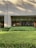 A building exterior with a facade made of light-colored bricks. Large, floor-to-ceiling glass windows line the wall, revealing an interior space that appears to be well-lit and furnished with elements like a lamp and framed art. In front of the building is a well-maintained garden with neatly trimmed bushes and greenery. Shadows from nearby trees are cast on the facade.