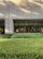 A building exterior with a facade made of light-colored bricks. Large, floor-to-ceiling glass windows line the wall, revealing an interior space that appears to be well-lit and furnished with elements like a lamp and framed art. In front of the building is a well-maintained garden with neatly trimmed bushes and greenery. Shadows from nearby trees are cast on the facade.