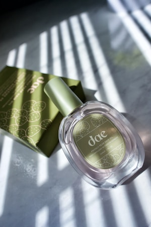 A bottle of prickly pear hair oil with a nearly transparent liquid is accompanied by a green box, both of which are placed on a surface with striped shadows cast over them. The packaging and bottle design feature a minimalist style with cactus illustrations.