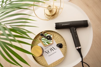 A stylish table arrangement features a modern black hairdryer labeled 'gama.professional'. The dryer is placed on a round white table next to a decorative gold tray holding a green glass bottle, a jar with a gold lid, a black crystal, and a small card with the word 'NOW'. A lush green palm leaf partially covers the table, adding a touch of nature.
