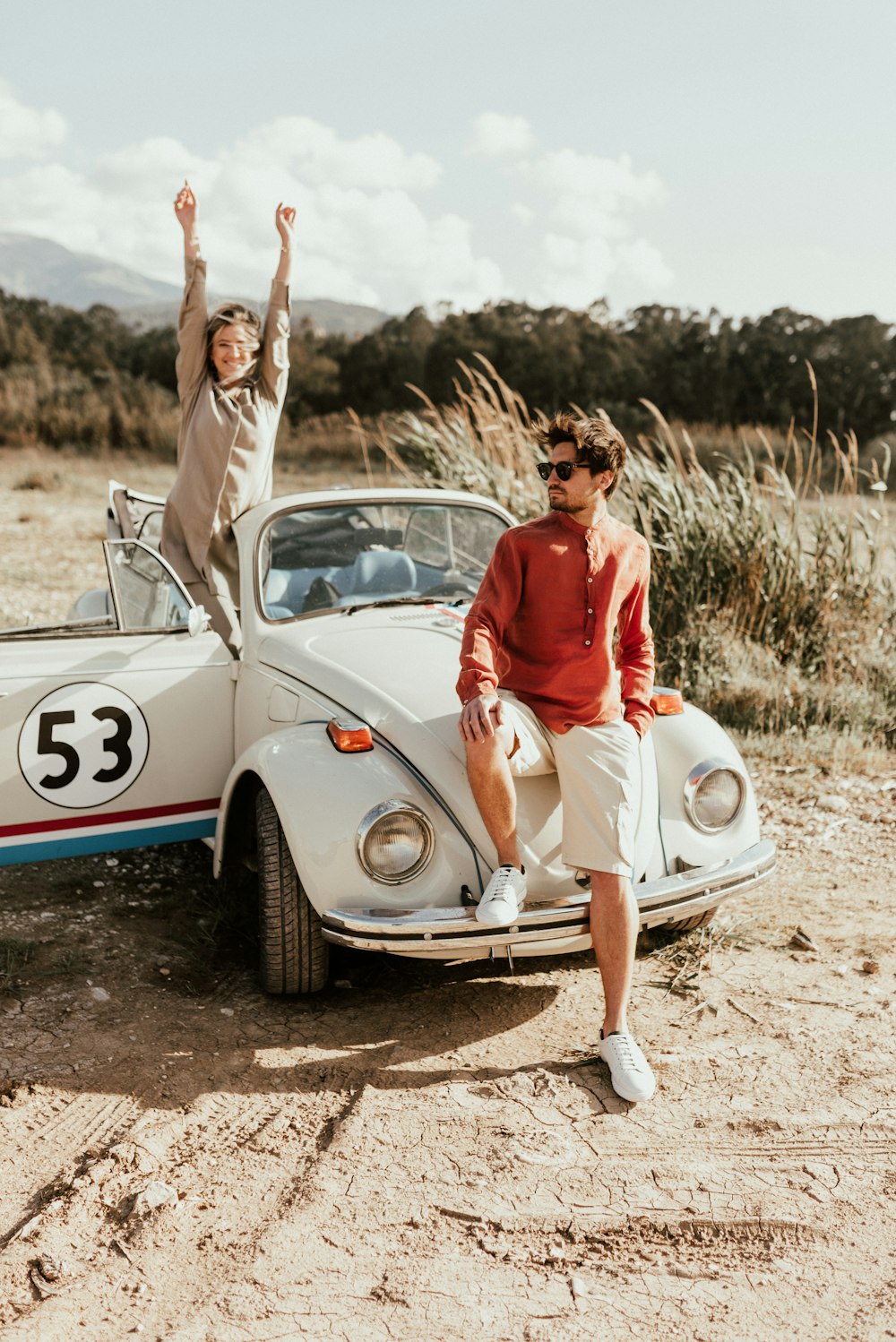 woman in red tank top and white pants sitting on white volkswagen beetle