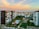 Numerous apartment buildings line a street, with balconies facing outwards. A tennis court and pool are visible in a residential complex. The backdrop consists of a distant cityscape and a coastline. The sky is filled with soft pastel hues as the sun sets.