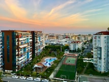 Numerous apartment buildings line a street, with balconies facing outwards. A tennis court and pool are visible in a residential complex. The backdrop consists of a distant cityscape and a coastline. The sky is filled with soft pastel hues as the sun sets.