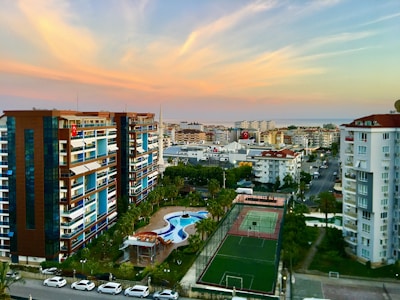 Numerous apartment buildings line a street, with balconies facing outwards. A tennis court and pool are visible in a residential complex. The backdrop consists of a distant cityscape and a coastline. The sky is filled with soft pastel hues as the sun sets.