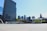 A spacious urban plaza featuring modern architecture with tall skyscrapers in the background and a large sign with colorful letters spelling 'TORONTO.' The area is open and appears well-lit with clear blue skies and some greenery visible.