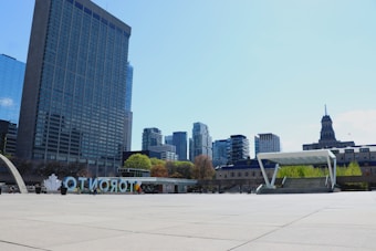 A spacious urban plaza featuring modern architecture with tall skyscrapers in the background and a large sign with colorful letters spelling 'TORONTO.' The area is open and appears well-lit with clear blue skies and some greenery visible.