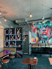 A trendy cafe interior features modern design elements like exposed light bulbs and a colorful abstract mural depicting a face. The walls include geometric neon lights in a tic-tac-toe pattern. There's a set of high tables and bar stools alongside cushioned seating.