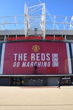 A large stadium facade with Manchester United written in prominent red letters. Below the name is a tall banner with the message 'The Reds Go Marching On!' in bold white text. A statue stands in front of the banner, and a person is seen walking near a megastore entrance.