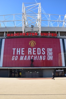 A large stadium facade with Manchester United written in prominent red letters. Below the name is a tall banner with the message 'The Reds Go Marching On!' in bold white text. A statue stands in front of the banner, and a person is seen walking near a megastore entrance.