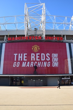 A large stadium facade with Manchester United written in prominent red letters. Below the name is a tall banner with the message 'The Reds Go Marching On!' in bold white text. A statue stands in front of the banner, and a person is seen walking near a megastore entrance.