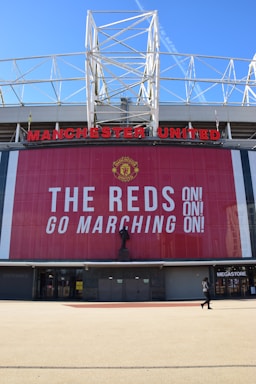 A large stadium facade with Manchester United written in prominent red letters. Below the name is a tall banner with the message 'The Reds Go Marching On!' in bold white text. A statue stands in front of the banner, and a person is seen walking near a megastore entrance.
