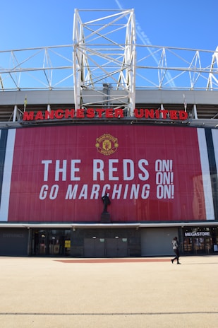 A large stadium facade with Manchester United written in prominent red letters. Below the name is a tall banner with the message 'The Reds Go Marching On!' in bold white text. A statue stands in front of the banner, and a person is seen walking near a megastore entrance.