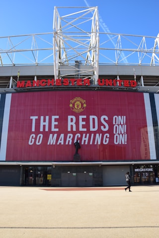 A large stadium facade with Manchester United written in prominent red letters. Below the name is a tall banner with the message 'The Reds Go Marching On!' in bold white text. A statue stands in front of the banner, and a person is seen walking near a megastore entrance.