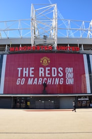 A large stadium facade with Manchester United written in prominent red letters. Below the name is a tall banner with the message 'The Reds Go Marching On!' in bold white text. A statue stands in front of the banner, and a person is seen walking near a megastore entrance.