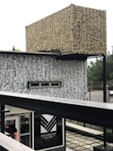An architectural structure with complex, abstract patterns covering its surfaces. The design is characterized by a combination of beige and black colors. It has a rectangular sign labeled 'Ventures Park' near the bottom with an entrance visible just below, where a person is partially visible. The background features some greenery with a slightly blurred appearance.