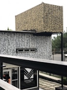 An architectural structure with complex, abstract patterns covering its surfaces. The design is characterized by a combination of beige and black colors. It has a rectangular sign labeled 'Ventures Park' near the bottom with an entrance visible just below, where a person is partially visible. The background features some greenery with a slightly blurred appearance.