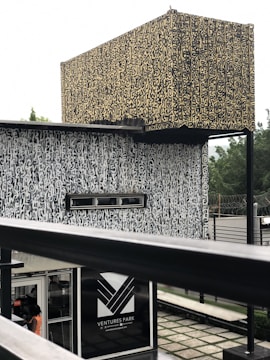 An architectural structure with complex, abstract patterns covering its surfaces. The design is characterized by a combination of beige and black colors. It has a rectangular sign labeled 'Ventures Park' near the bottom with an entrance visible just below, where a person is partially visible. The background features some greenery with a slightly blurred appearance.